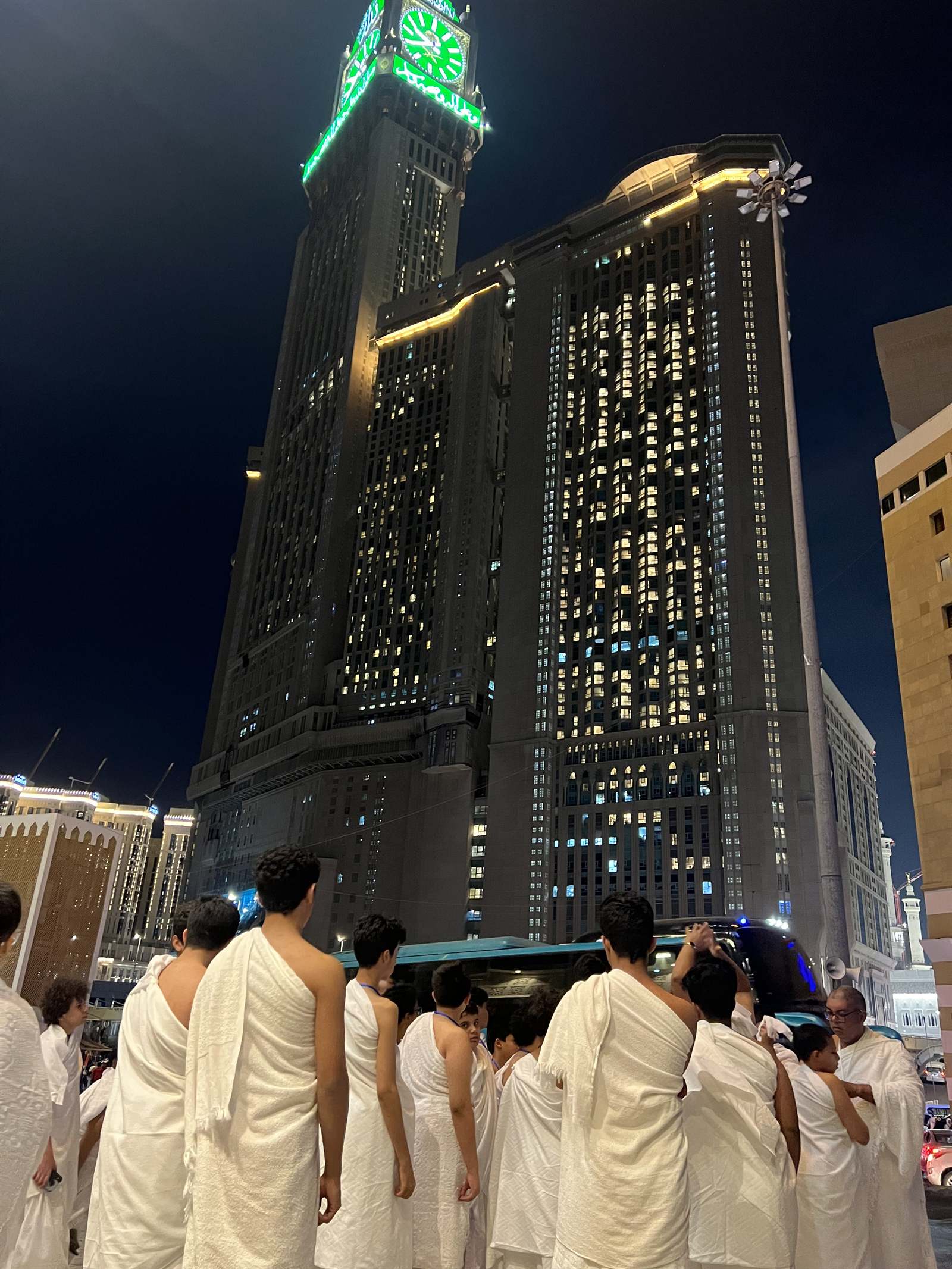 A group in ihram walking toward the Haram and the Clock Tower at night